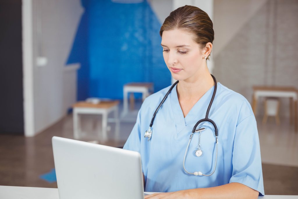 Female nurse wearing scrubs typing on laptop at desk in reception area with stethoscope, copy space. Medical staff, healthcare, professionalism, clinical, modern interior, patient care, wellness